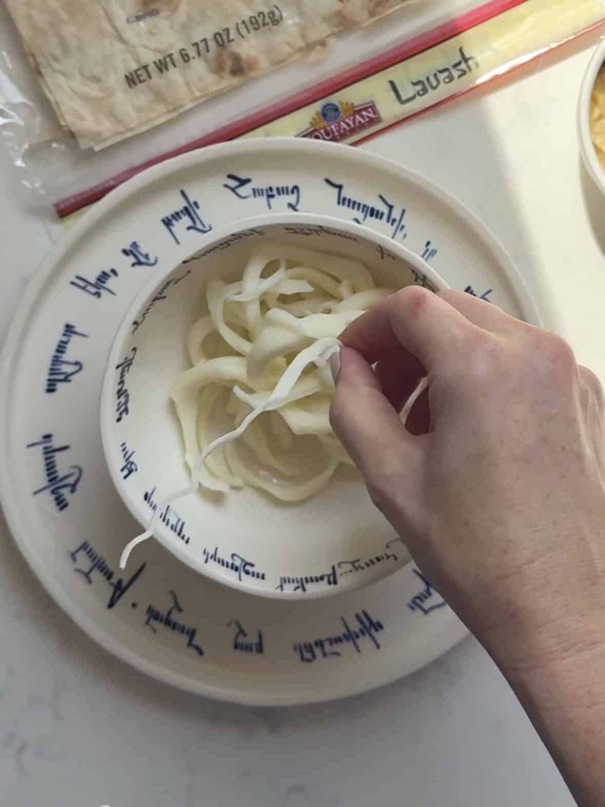 Hand pulling Armenian string cheese into thin strands in a bowl next to a package of Toufayan lavash.