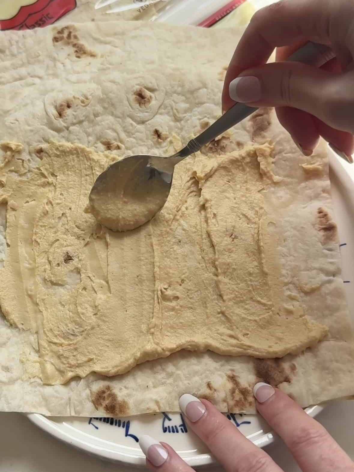 Spreading a thin layer of hummus over a sheet of lavash with a spoon.
