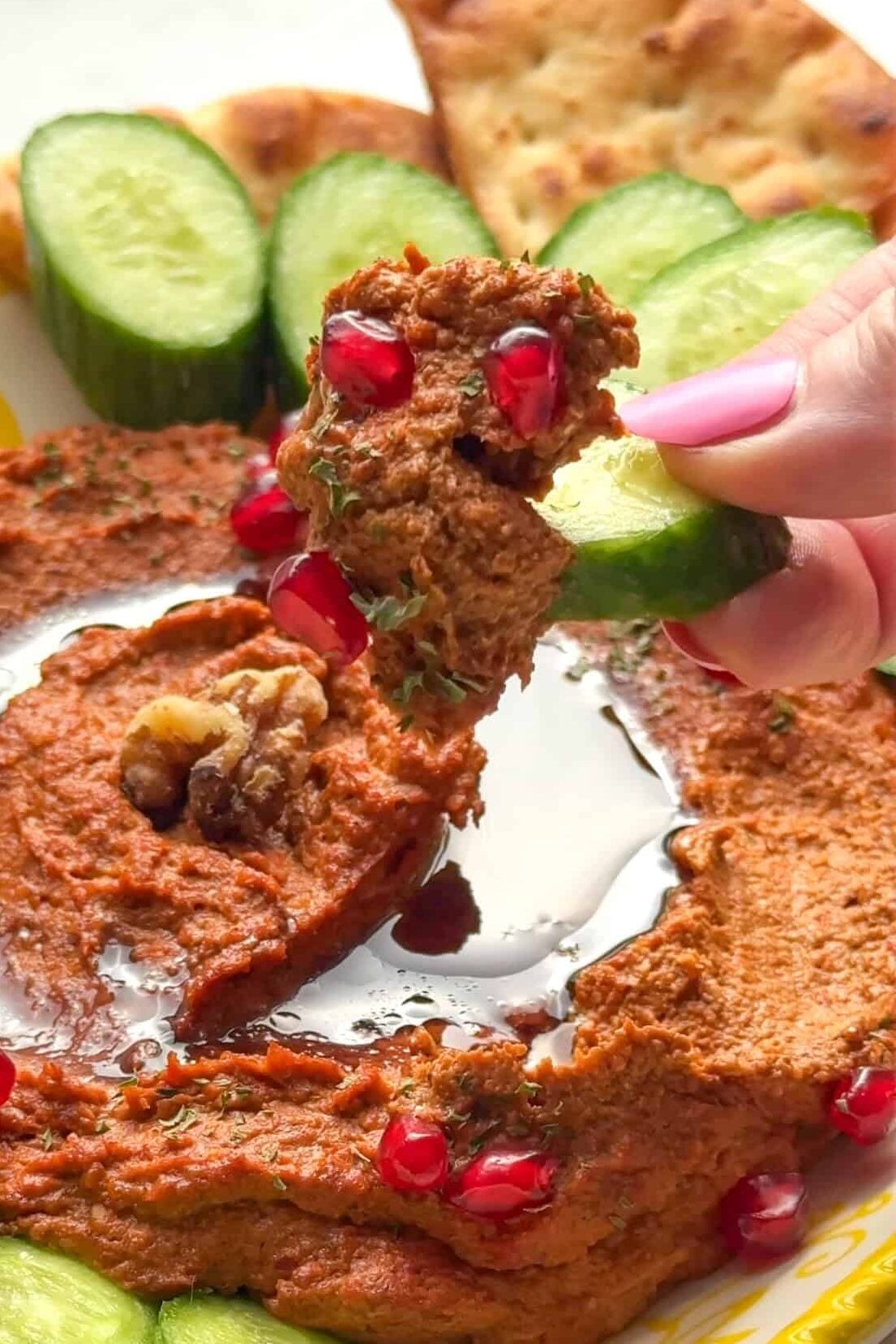 A hand holding pita triangles next to a platter of muhammara topped with pomegranate seeds and pomegranate molasses.