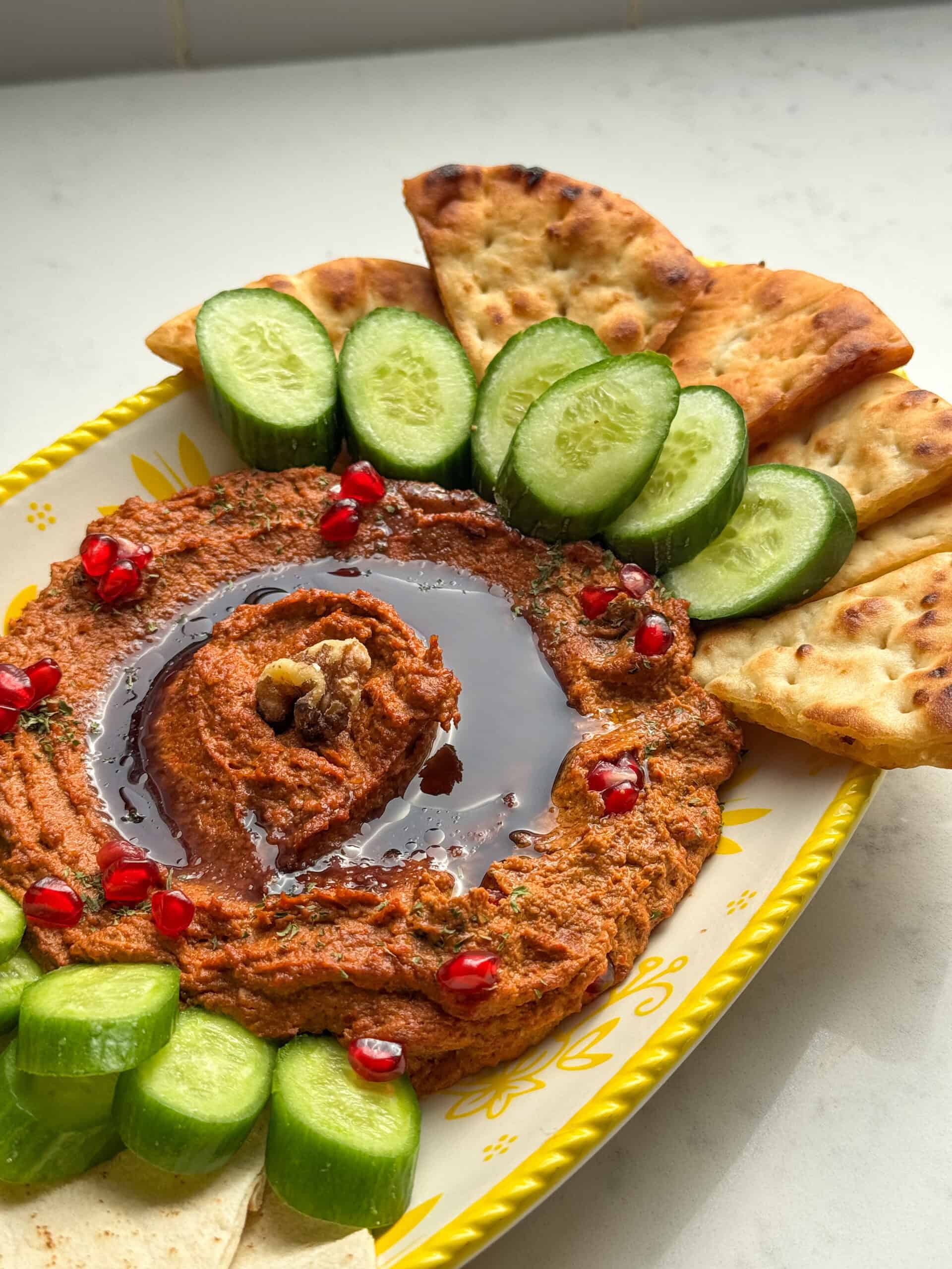 A platter of muhammara dip with toasted naan triangles and sliced cucumbers on a yellow and white plate.