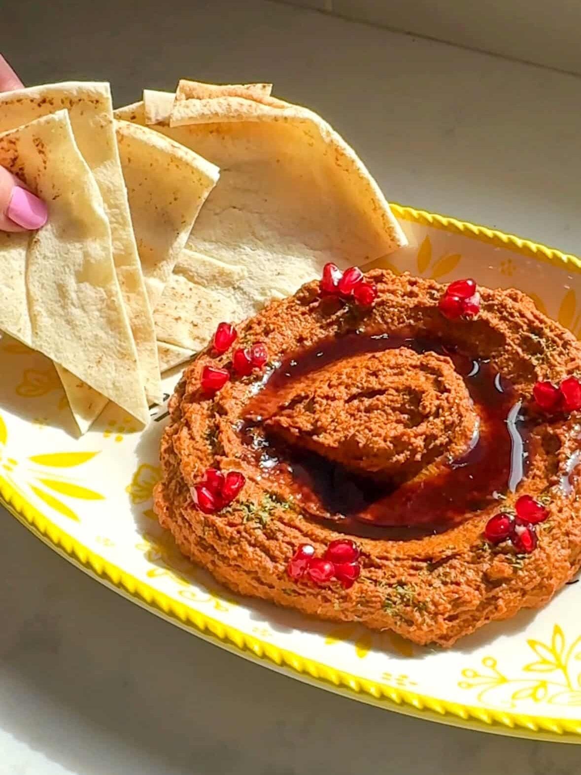 A hand holding pita triangles next to a platter of muhammara topped with pomegranate seeds and pomegranate molasses.