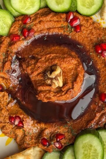 Overhead view of muhammara swirled with pomegranate molasses and topped with pomegranate seeds, surrounded by cucumbers, pita, and naan.