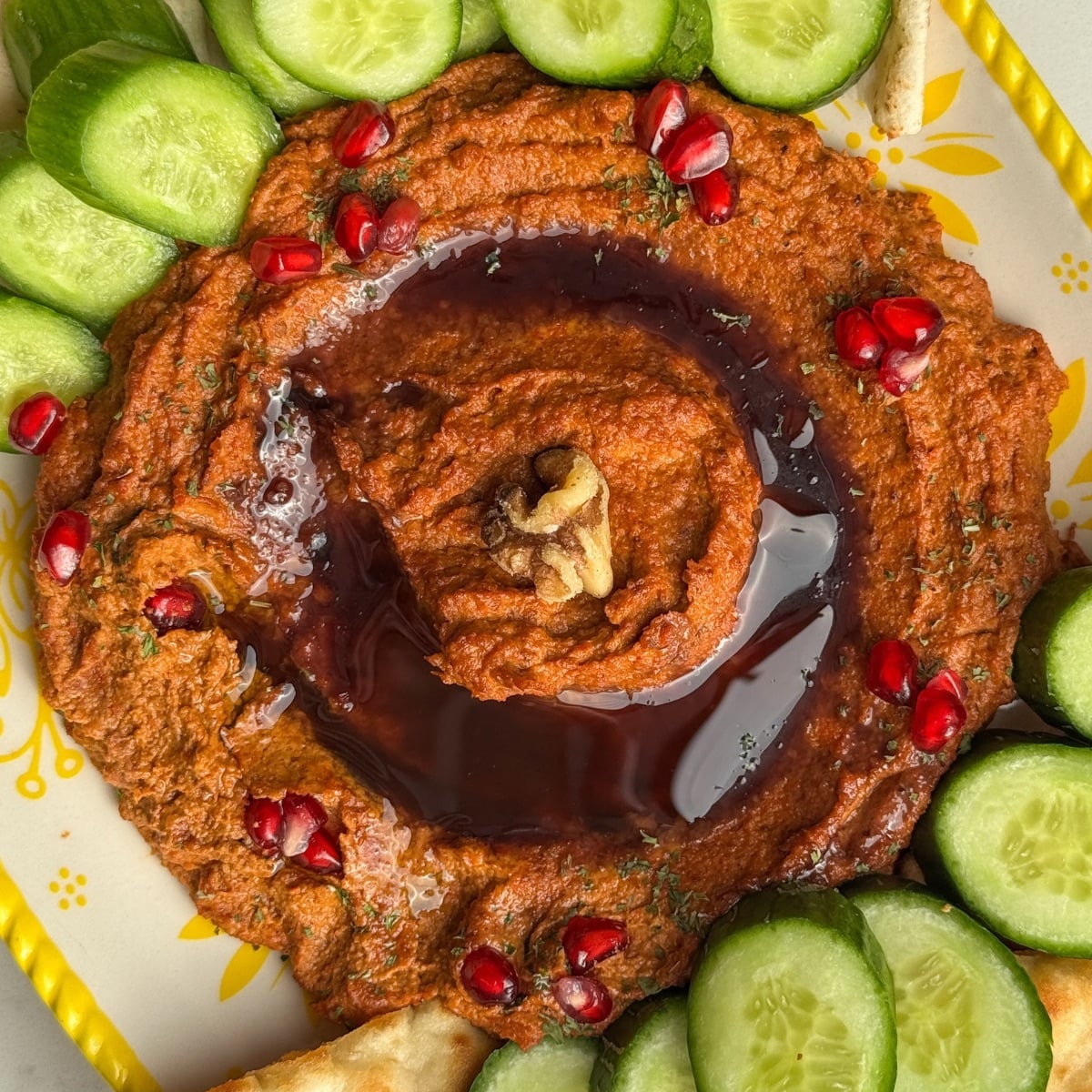 Overhead view of muhammara swirled with pomegranate molasses and topped with pomegranate seeds, surrounded by cucumbers, pita, and naan.
