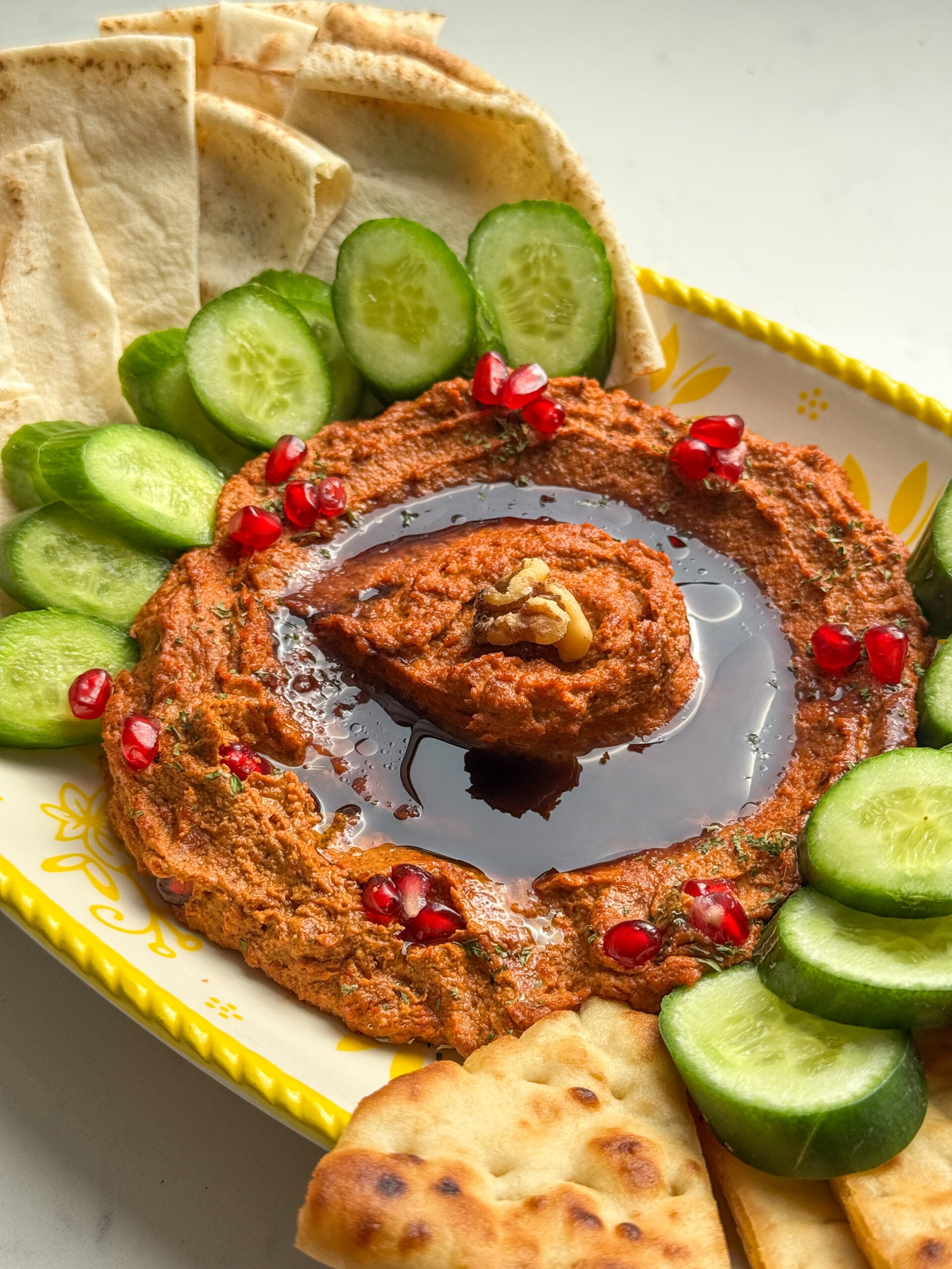 Muhammara roasted red pepper and walnut dip with pomegranate molasses, pomegranate seeds, and a walnut, served with cucumbers, pita, and naan.