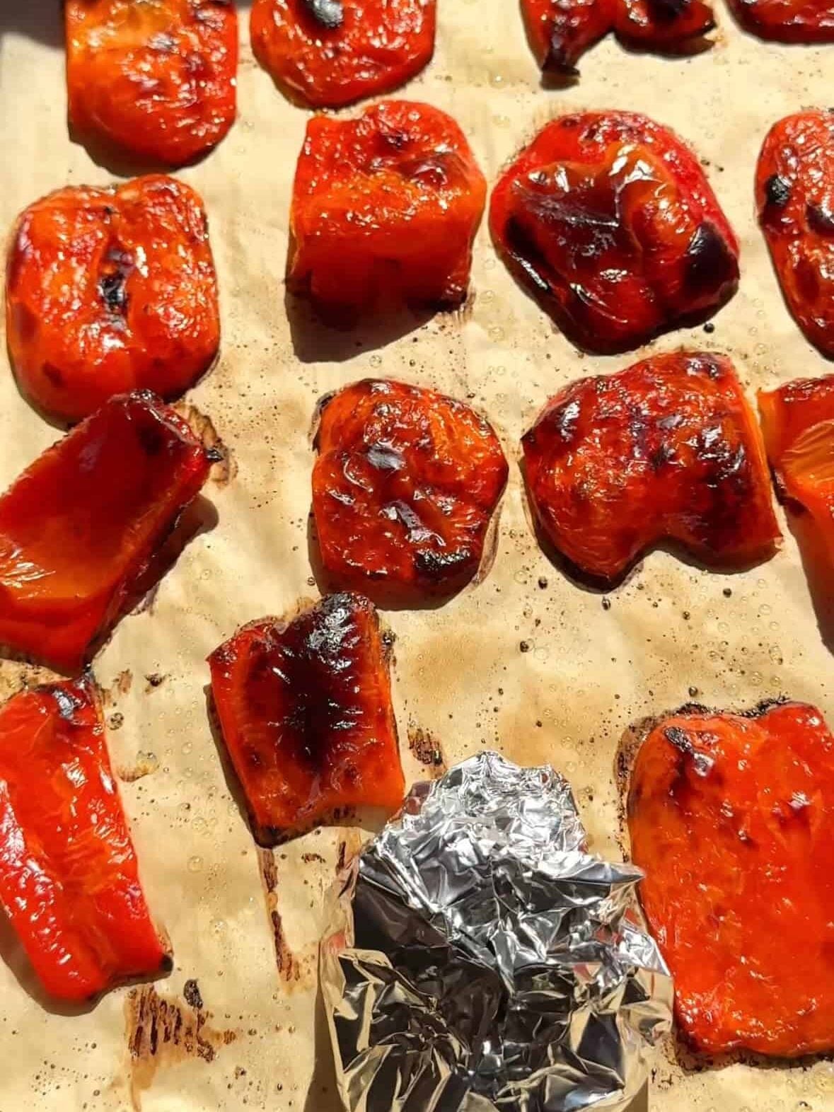 Roasted and charred red bell peppers on a parchment-lined baking sheet with a foil-wrapped head of garlic.