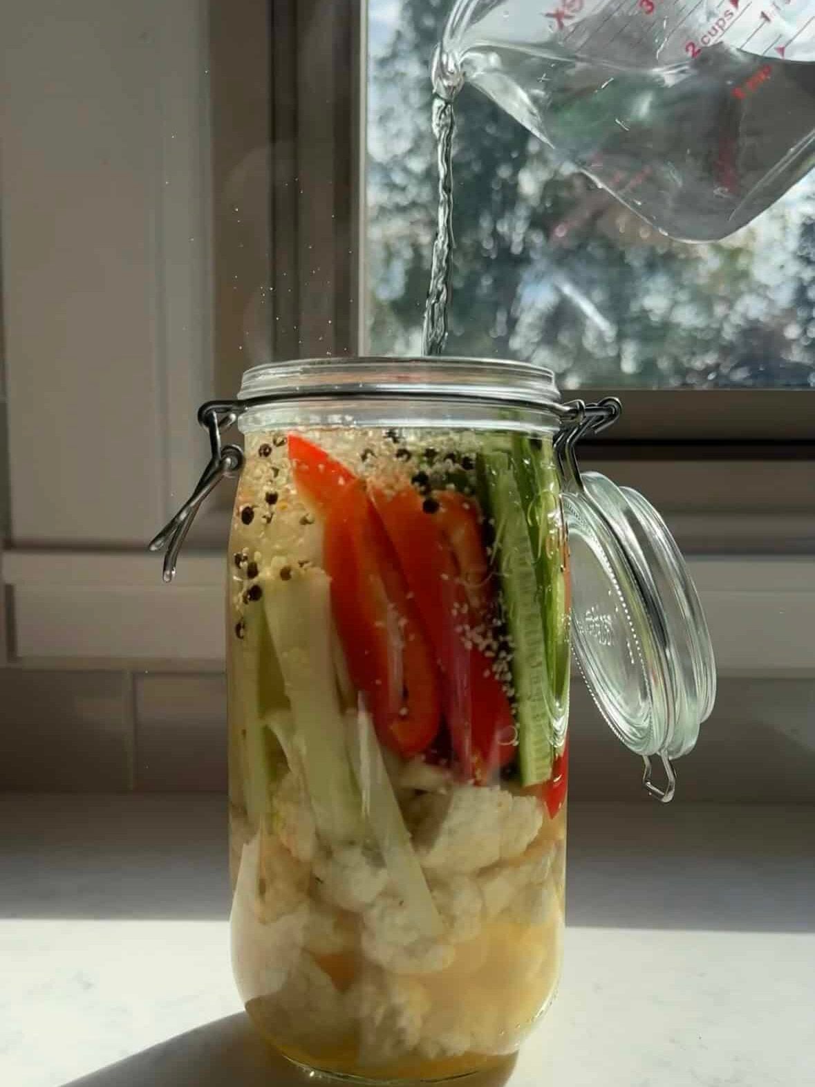 Pouring salt and vinegar brine into a jar of vegetables with black peppercorns and garlic