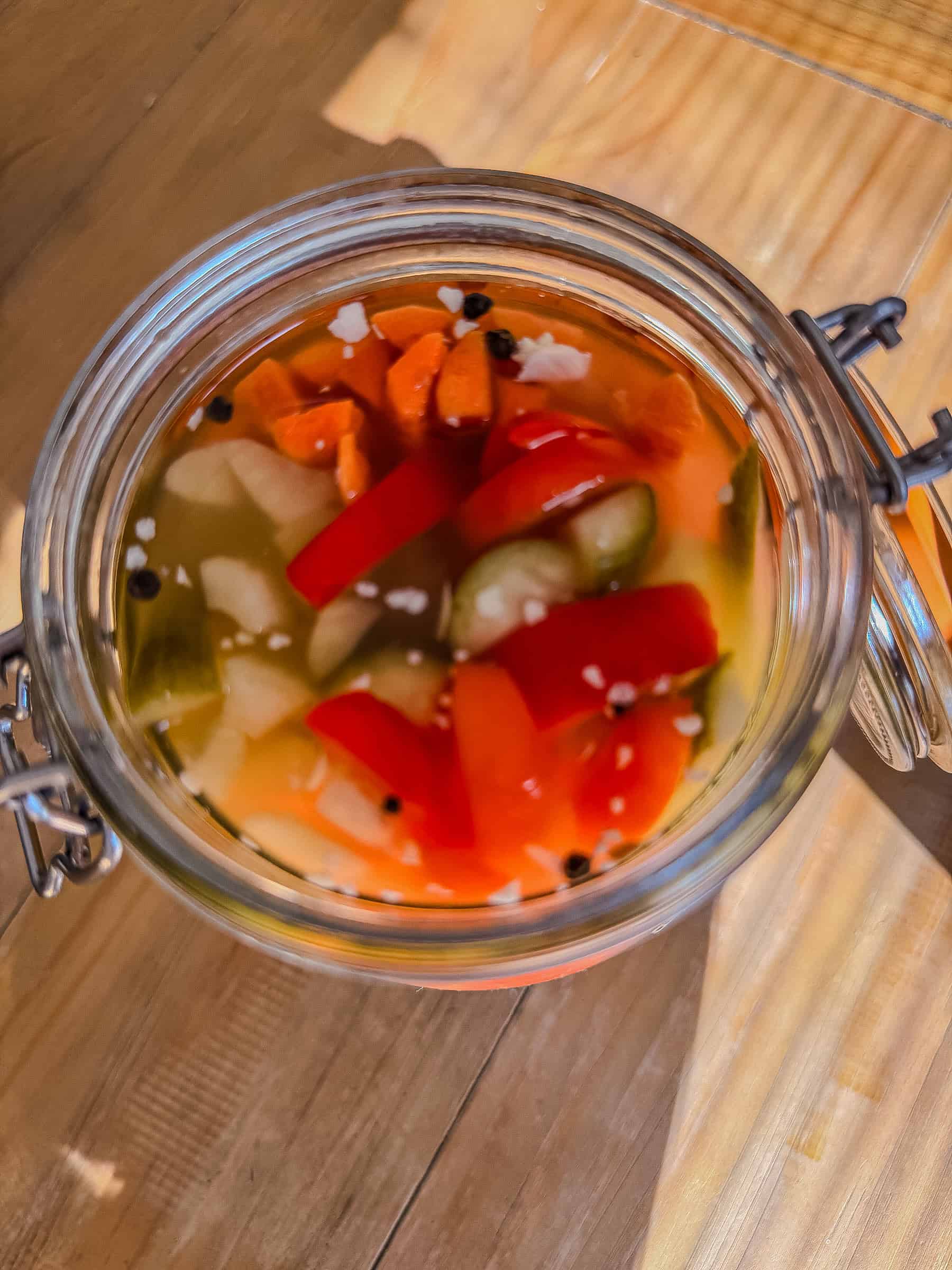 Overhead view of tourshi in an open jar with red pepper, carrots, cucumber, garlic, and peppercorns in brine